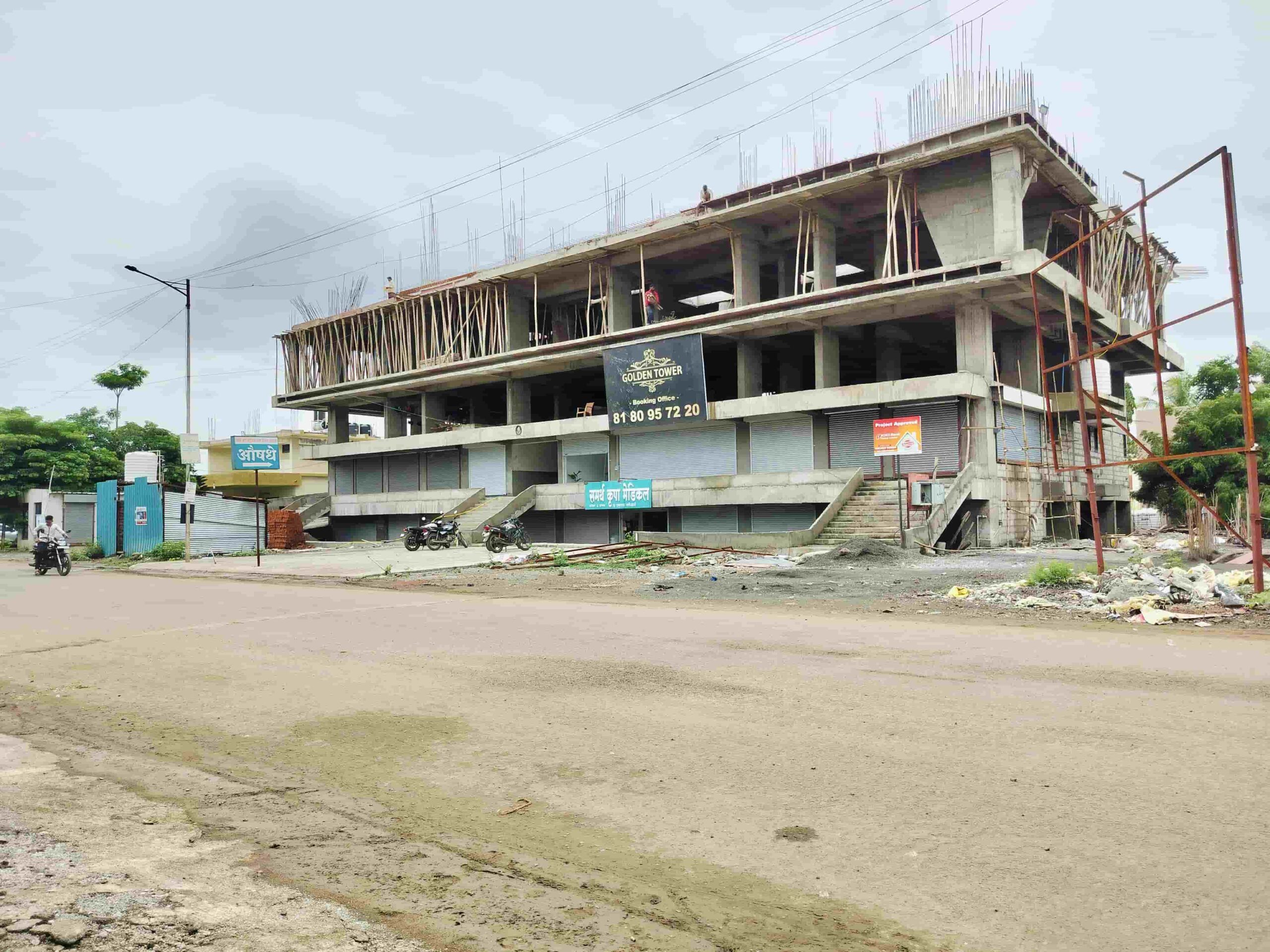 This image shows a building under construction on the side of a road. The building is made of concrete and steel, and it is surrounded by scaffolding. There are several workers on the site, and they are busy working on the building. The overall atmosphere is busy and productive.